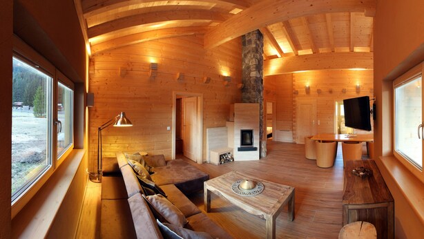 Interior de una cabaña de troncos con techo de madera, chimenea de piedra, sofá, mesa de centro y televisor.