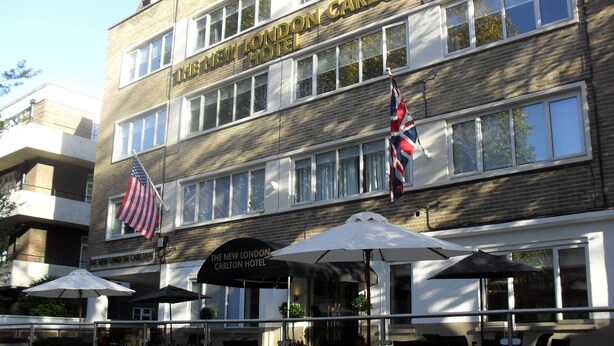 Het uiterlijk van het New London Carlton Hotel met een twee verdiepingen tellende bakstenen gevel, vlaggen van Amerika en de Union Jack, en terrasstoelen met parasols.