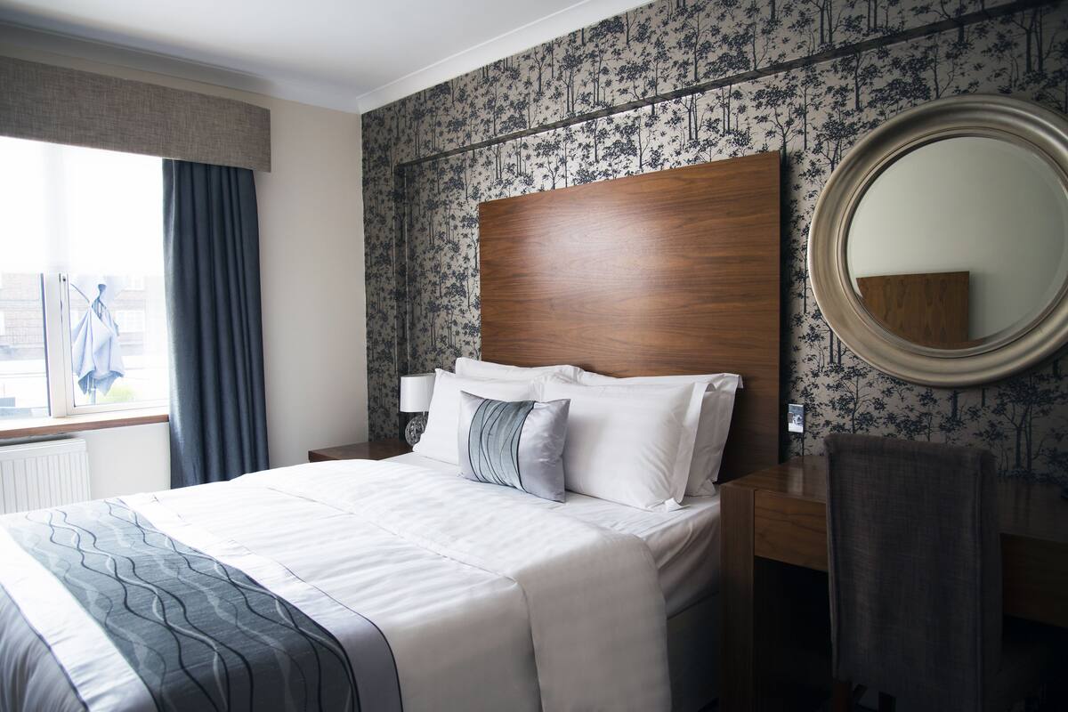 A neatly made bed with a wooden headboard, a round mirror, and a nightstand with a lamp.