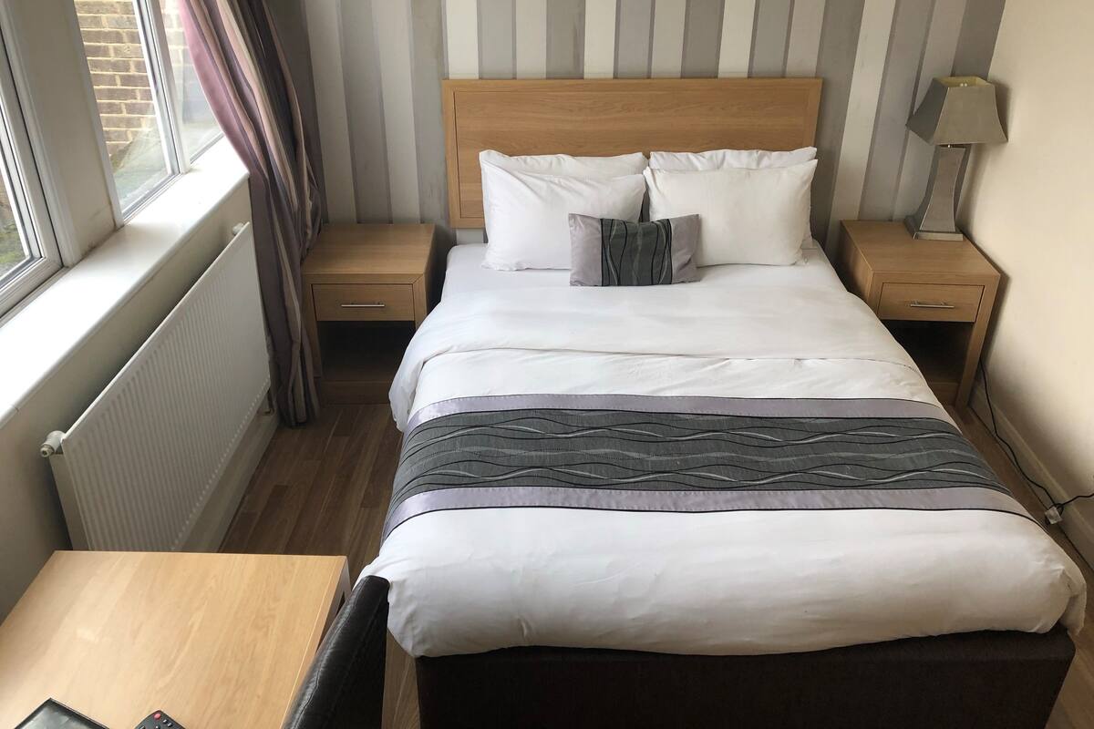 A hotel room with a wooden headboard, white bedding, a bedside table, a lamp, and a view of the outside through a window.
