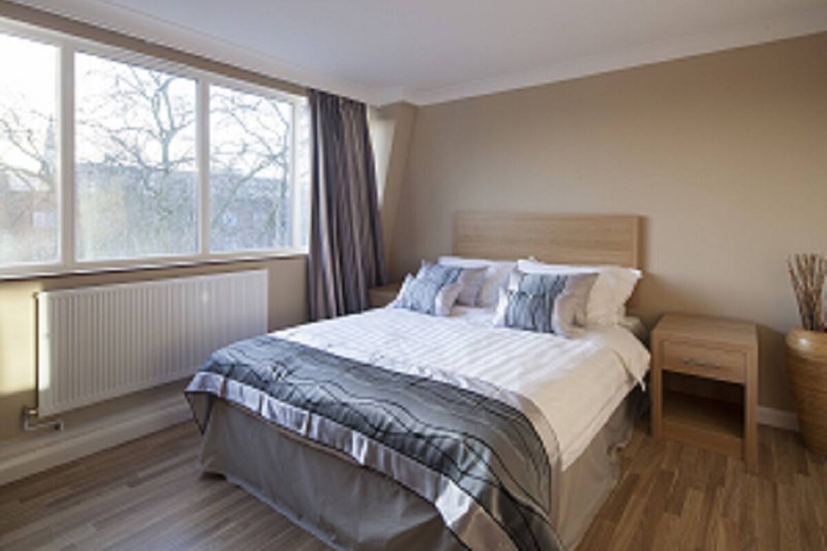 A bedroom with a large bed, a nightstand, a window with curtains, and a radiator.