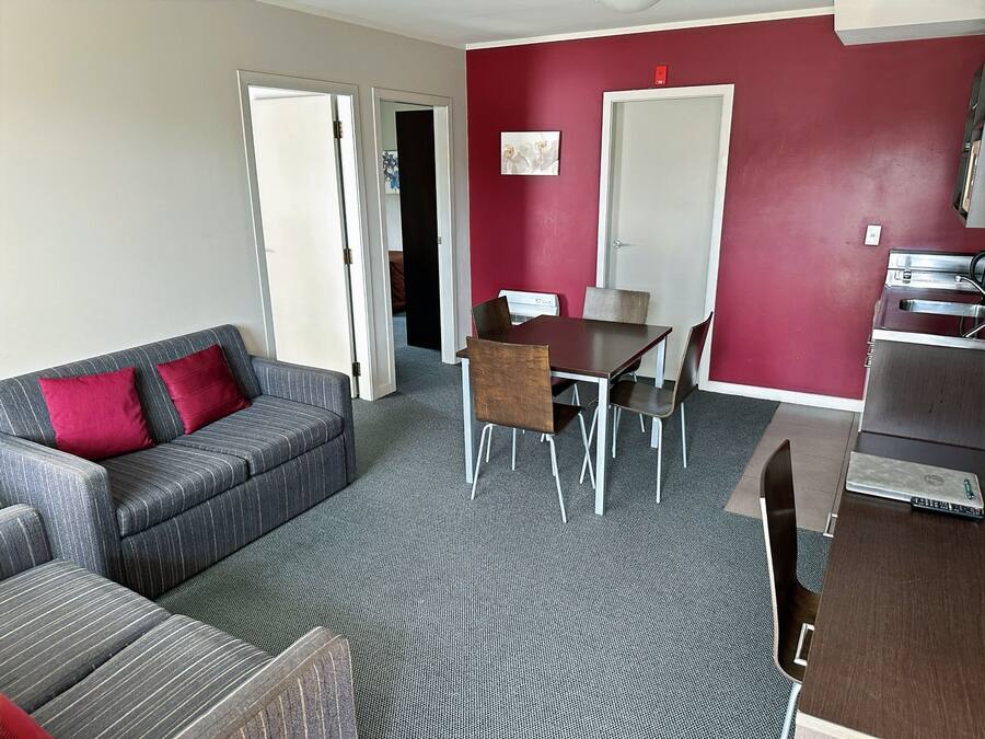 Un espacio habitable compacto con sofá gris, cojines rojos, una mesa de comedor con sillas, una kitchenette y una pared de acento roja.