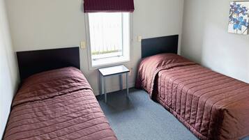 A small, clean room with two single beds, a window with a curtain, a small table, and a painting on the wall.