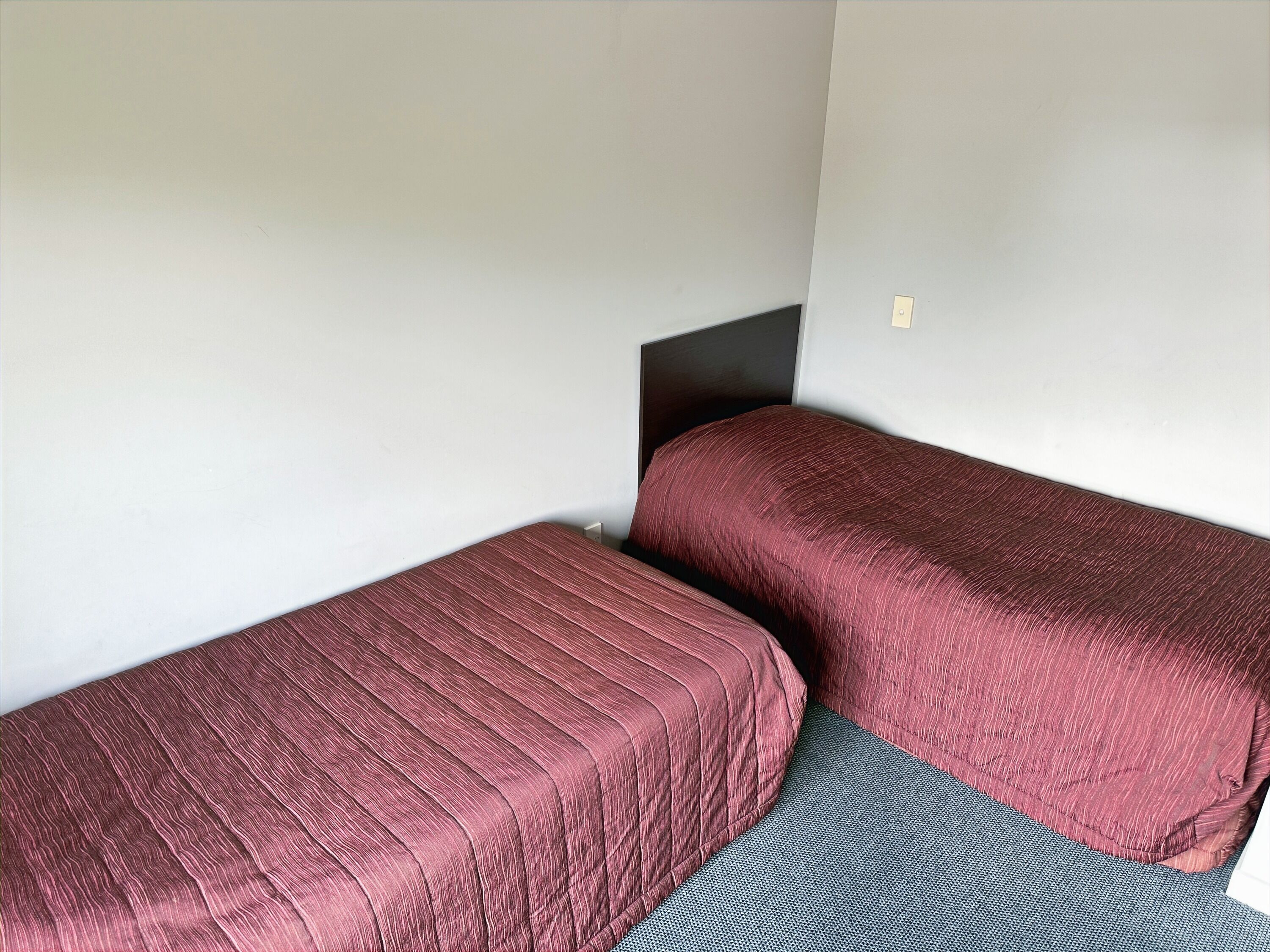 A small, single-room accommodation with two single beds, a dark headboard, and a plain white wall.