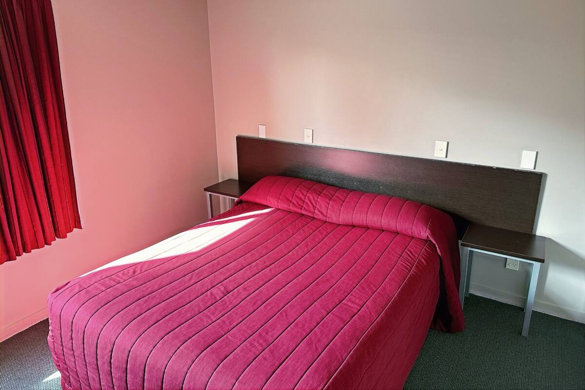 Habitación de hotel con cama, mesita de noche, cortina roja y pared blanca.
