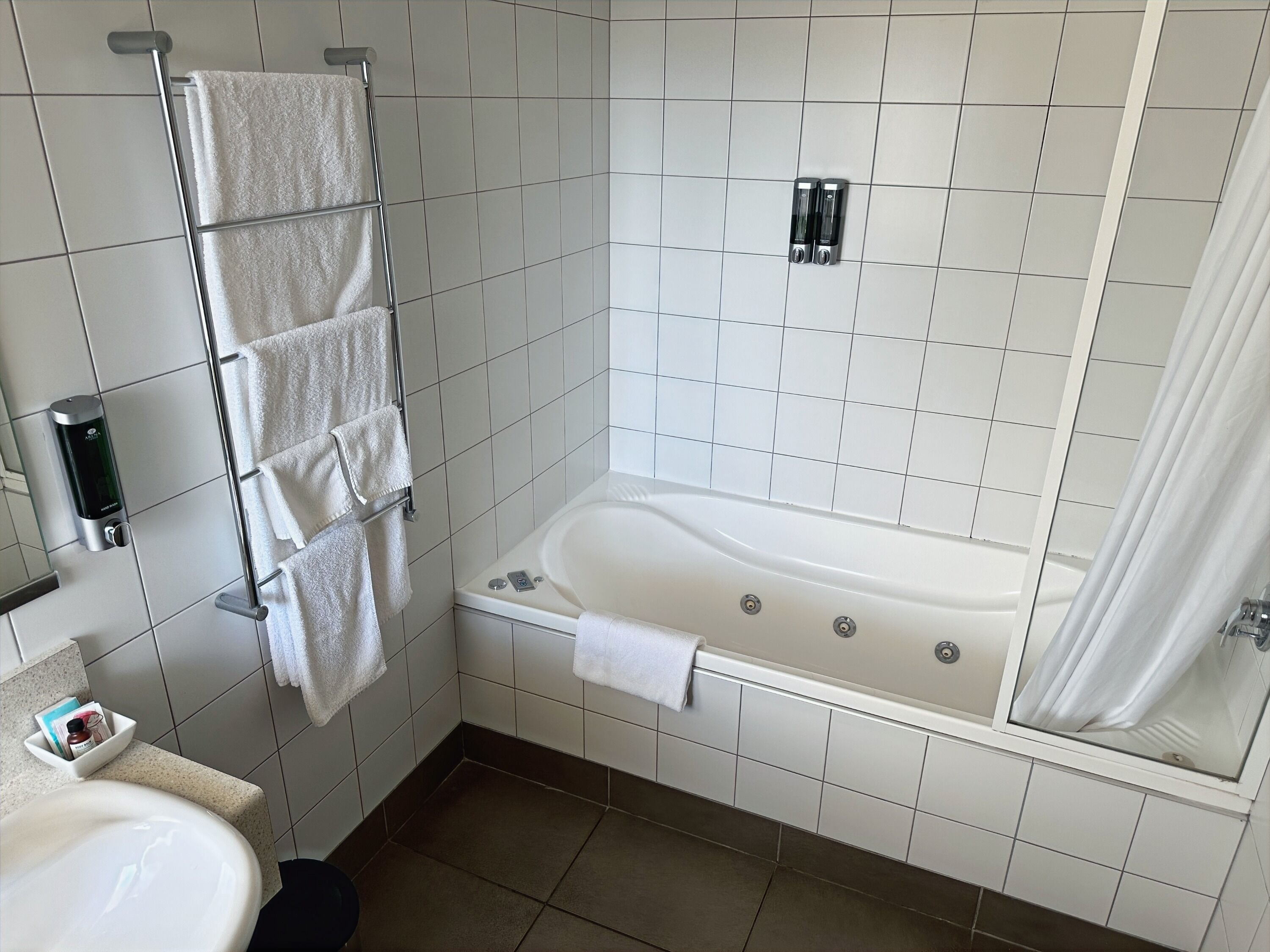 A modern bathroom with a white bathtub, a towel rack with multiple towels, a soap dispenser, and a shower curtain.