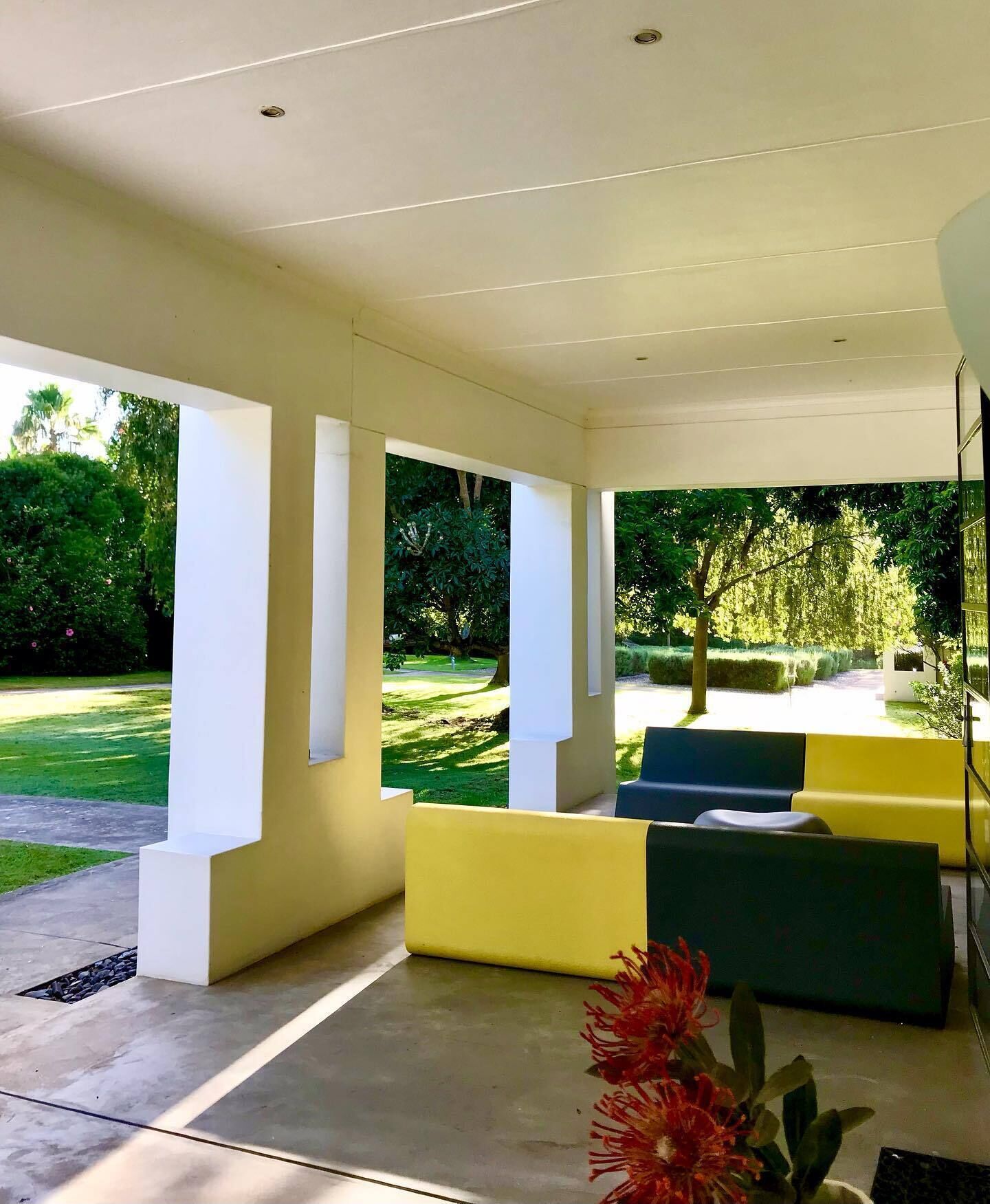 A modern outdoor seating area with yellow and green sofas, a concrete floor, and a view of a well-maintained garden.