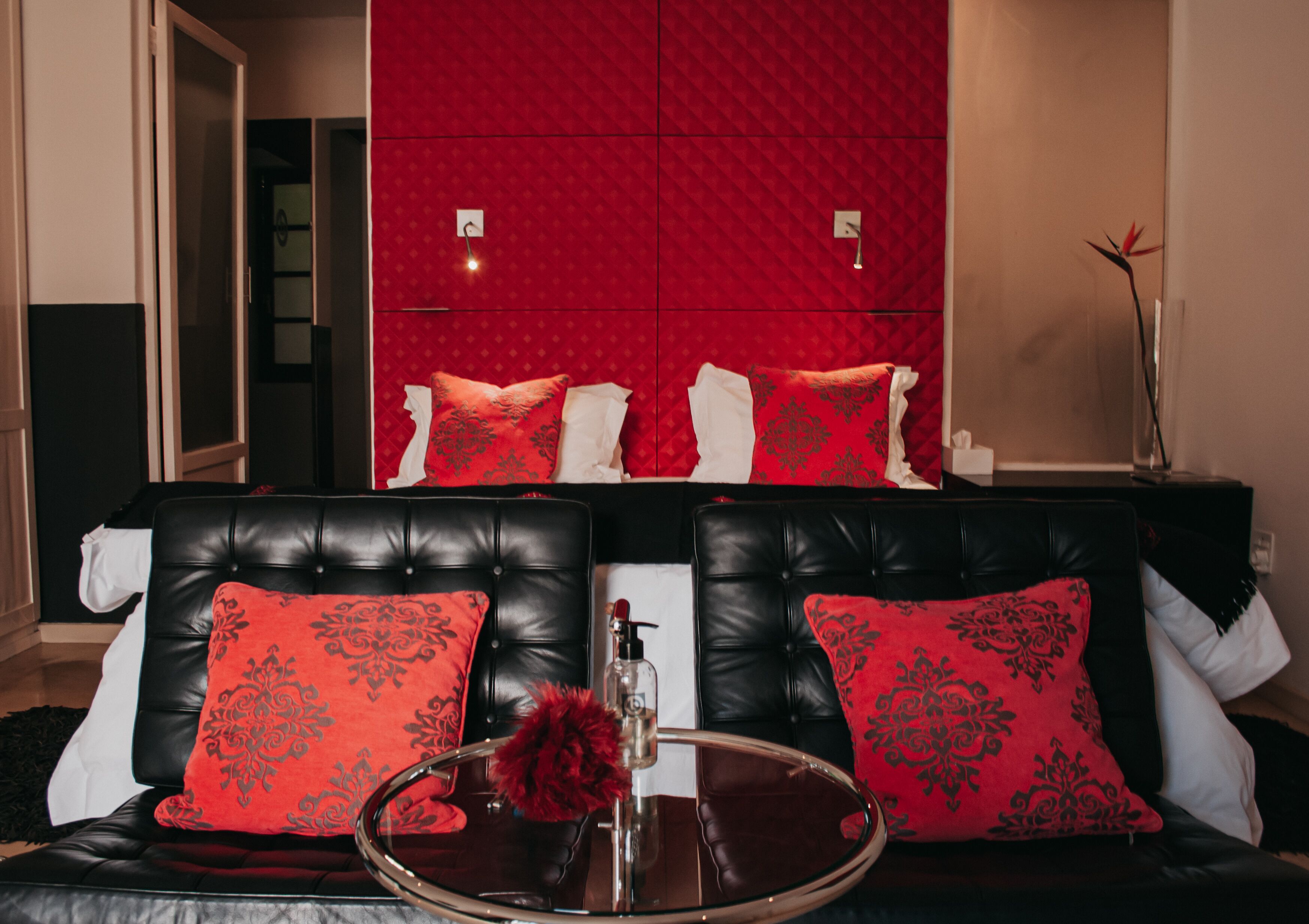Une pièce avec un canapé en cuir noir, des coussins aux motifs rouges et blancs, une table basse en verre ornée d’un objet décoratif rouge, et un mur rouge à texture derrière le canapé.