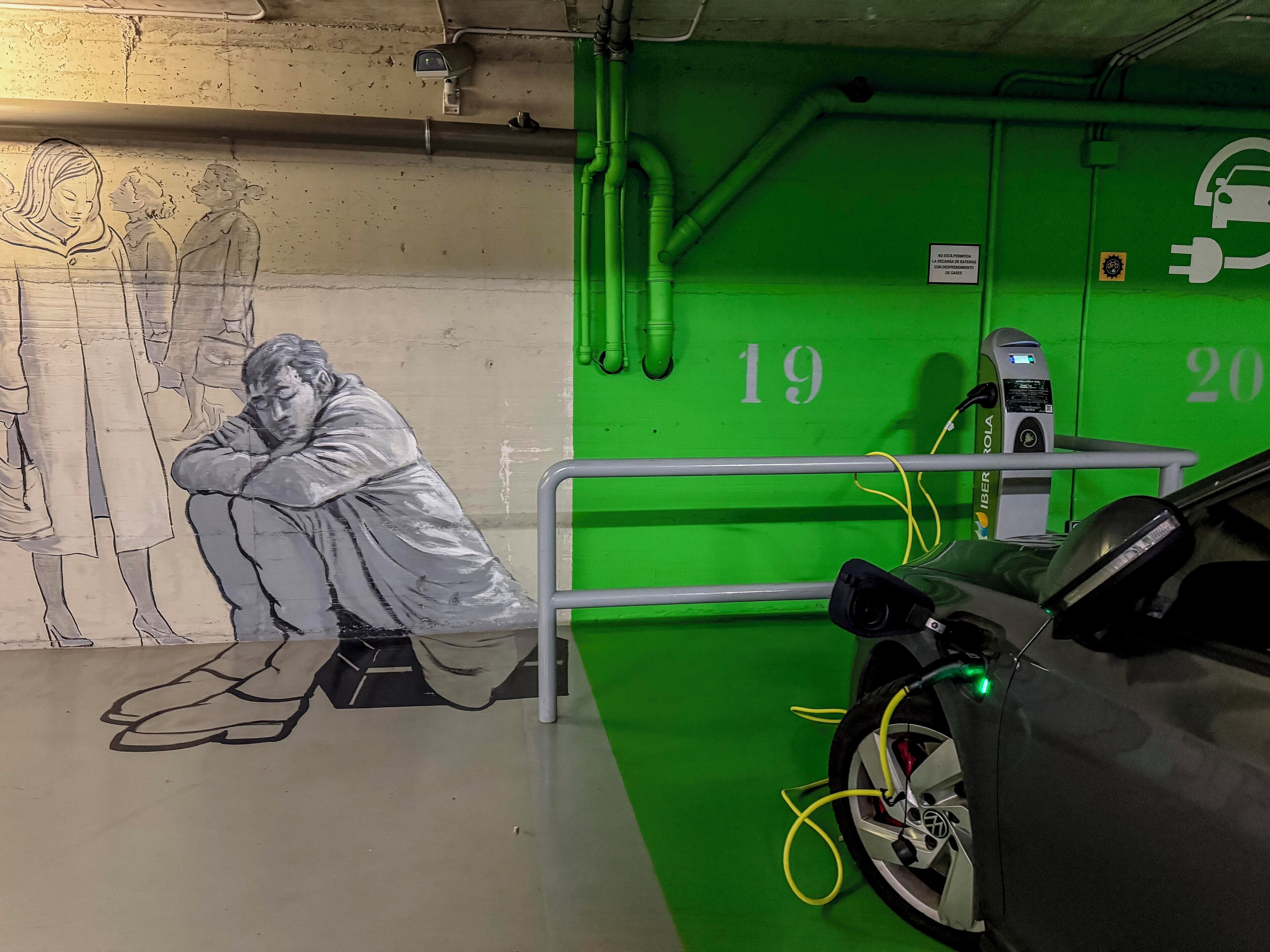 Un aparcamiento con paredes verdes, plazas numeradas y una estación de carga para vehículos eléctricos.