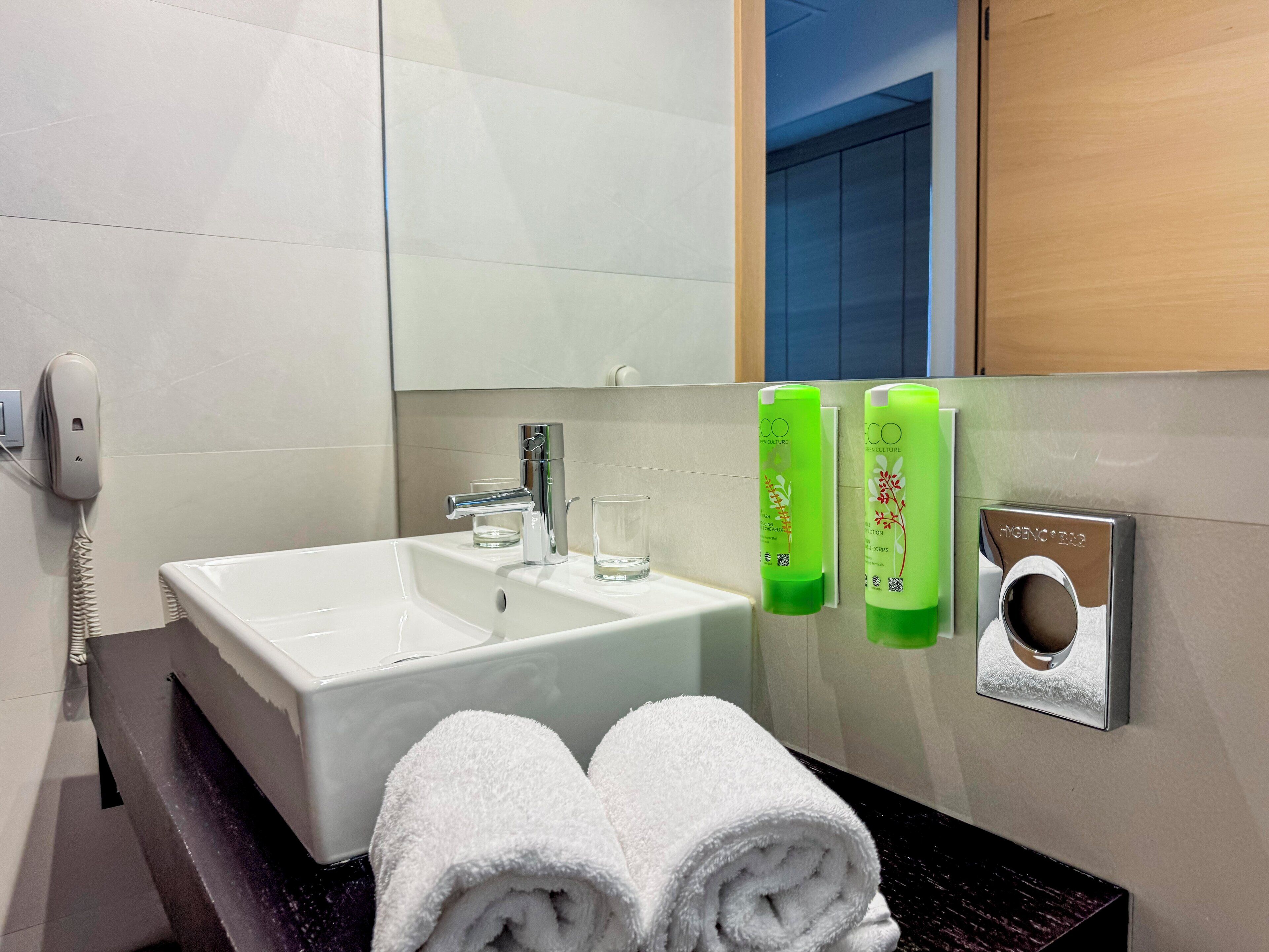 Un bany d'hotel amb un rentapeus blanc, un mirall, dues ampolles verdes i dues tovelloles blanques.