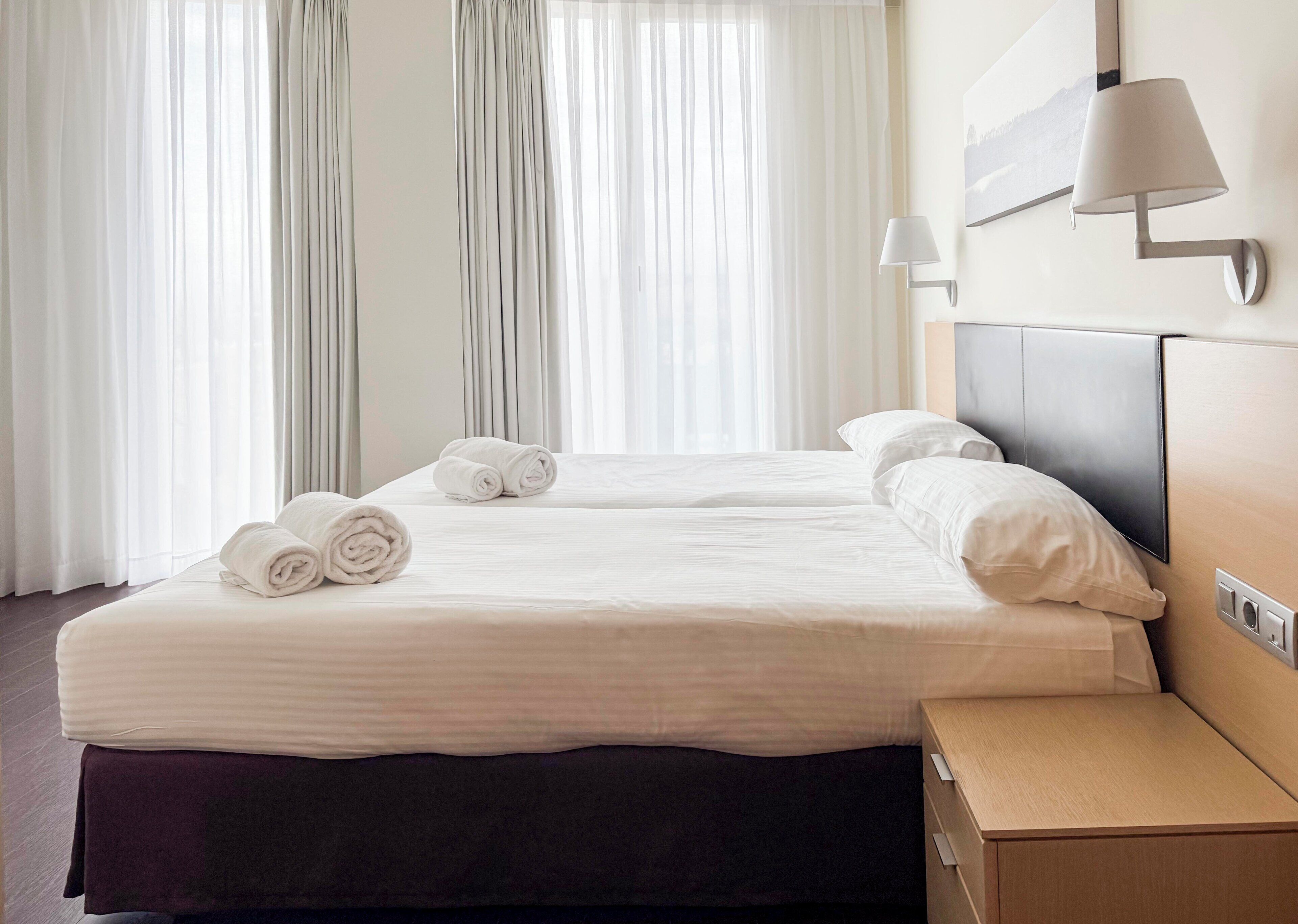 Habitación de hotel con dos camas, una mesita de noche, una lámpara y un ventanal con cortinas translúcidas.