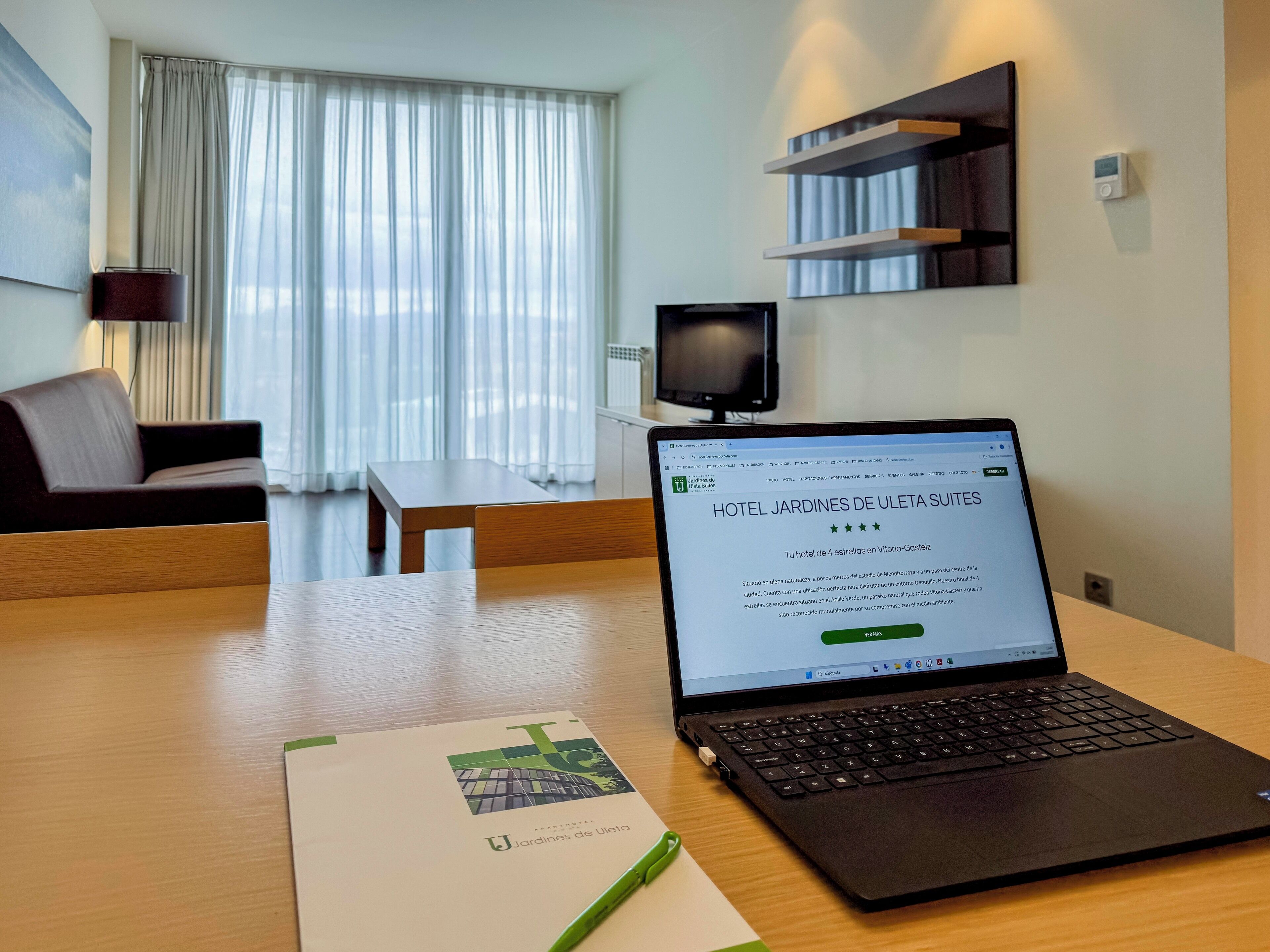 En bærbar computer på et træbord med en hotelbrochure og en pen. Et fjernsyn er monteret på væggen, og en sofa ses i baggrunden.