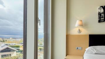 Una habitación de hotel con un ventanal grande, una cama, una mesita de noche y una lámpara fijada a la pared.