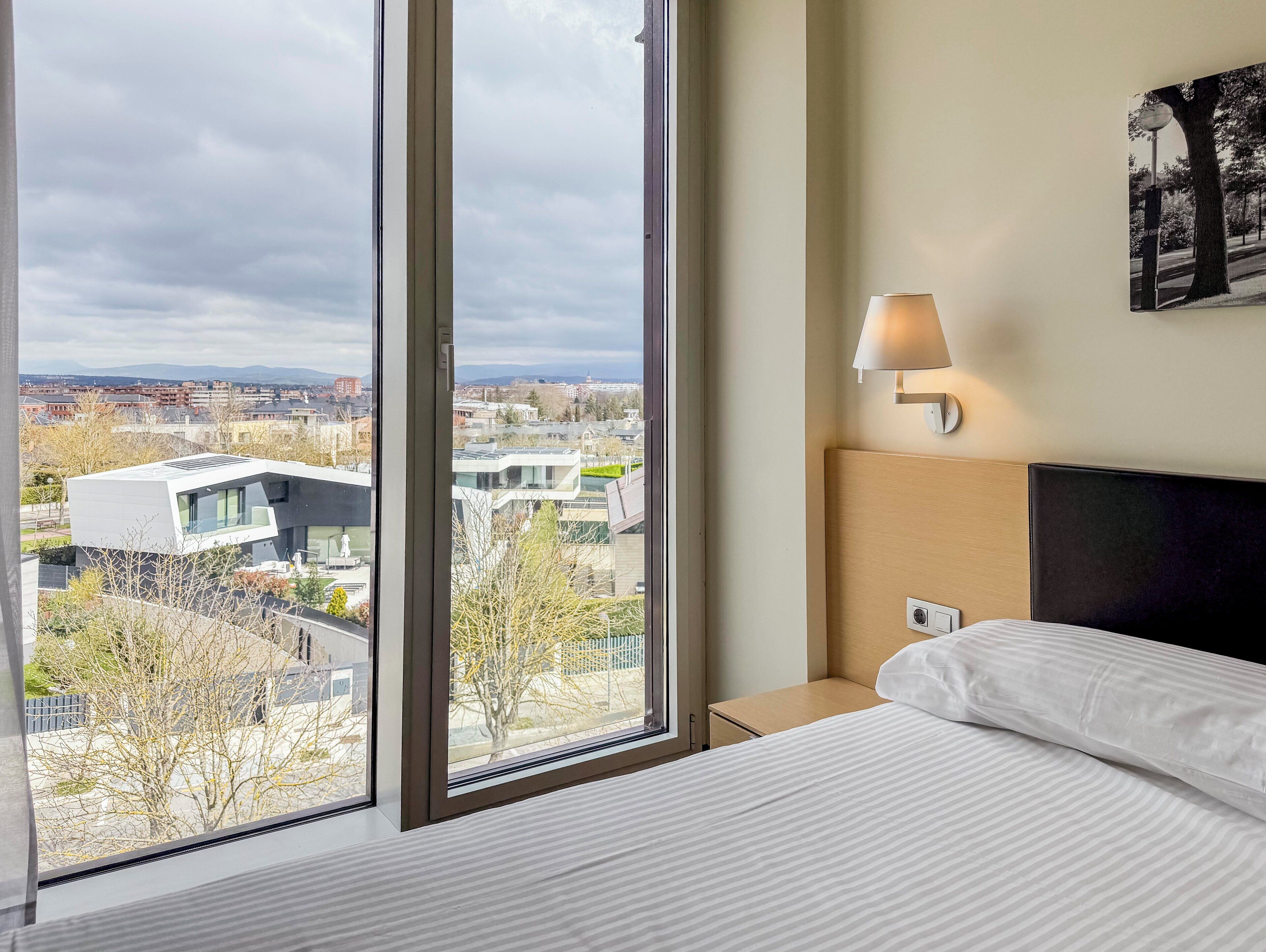 Habitación de hotel con un ventanal amplio que ofrece vistas a la ciudad, una cama con ropa blanca, una mesita de noche y una lámpara fijada a la pared.