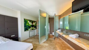 Una habitación de hotel moderna con una cama grande, ducha envuelta en cristal, tocador con espejo y un cuadro en la pared.