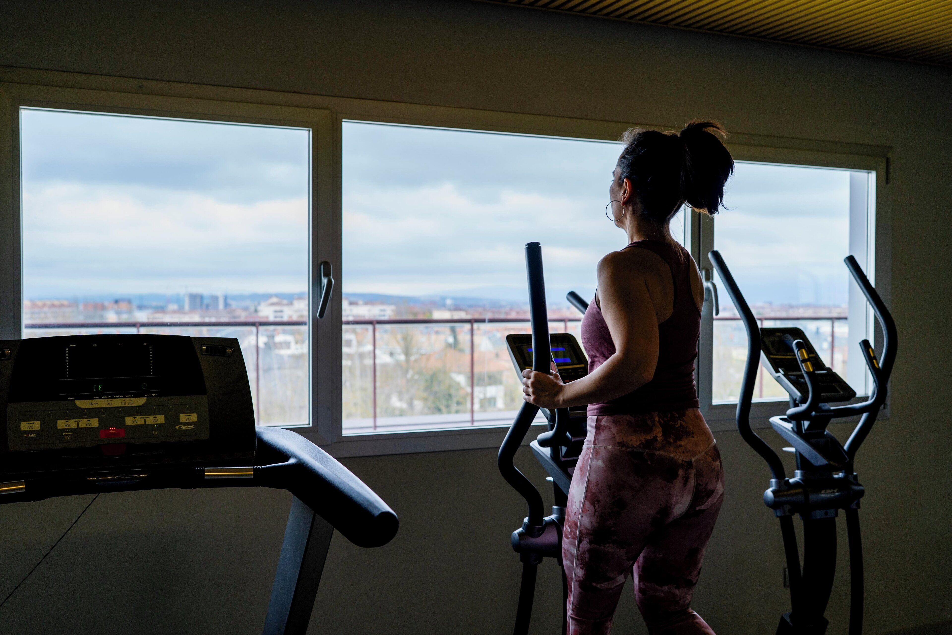 A gym with a treadmill and an elliptical machine, a large window with a city view, and a control panel on the treadmill.