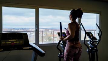 A gym with a treadmill and an elliptical machine, a large window with a city view, and a control panel on the treadmill.