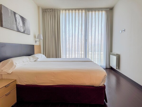 Habitación de hotel con dos camas, un ventanal con cortinas, un radiador y un cuadro en la pared.