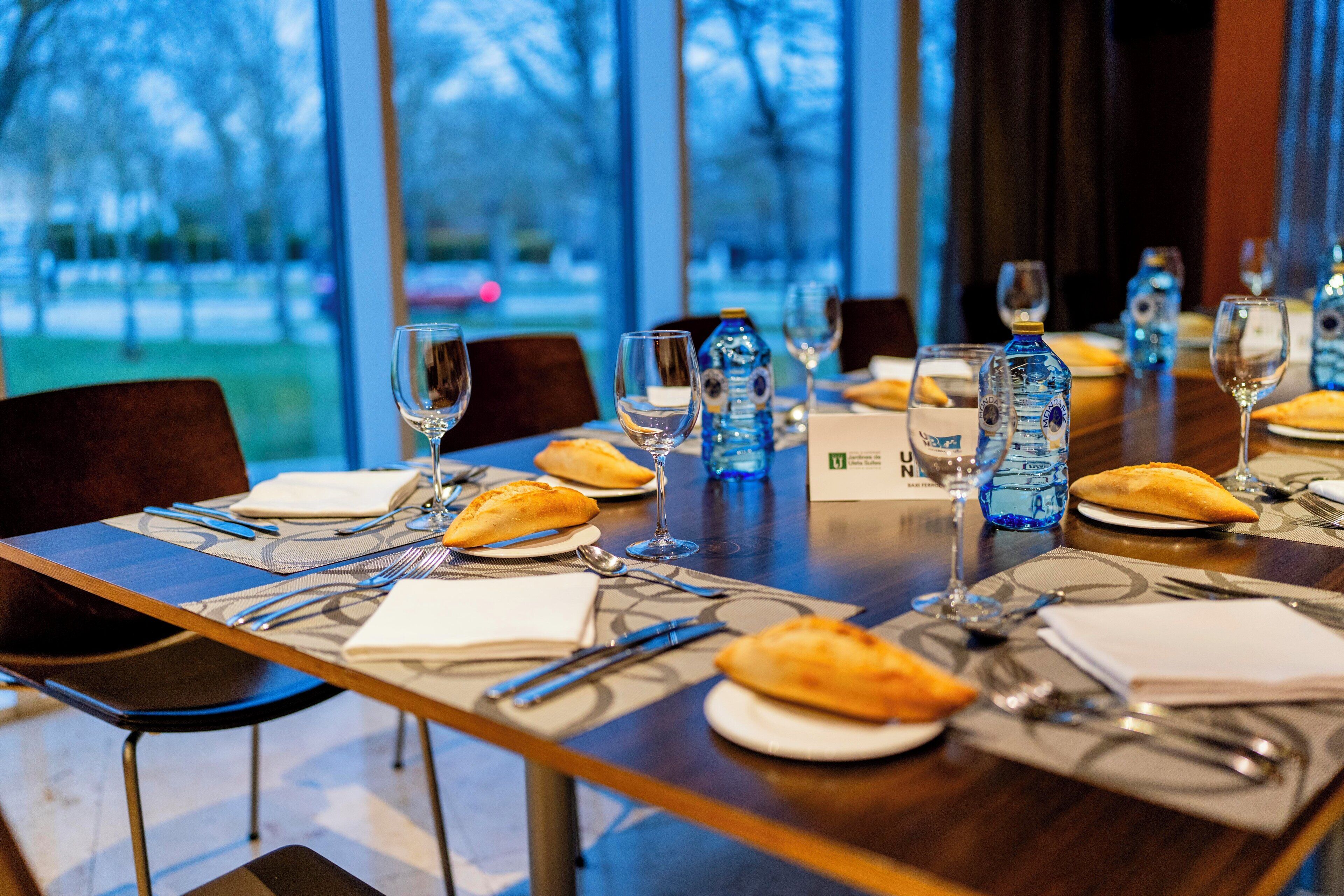 A dining table set with plates, cutlery, glasses, and water bottles, with a view of trees through large windows.