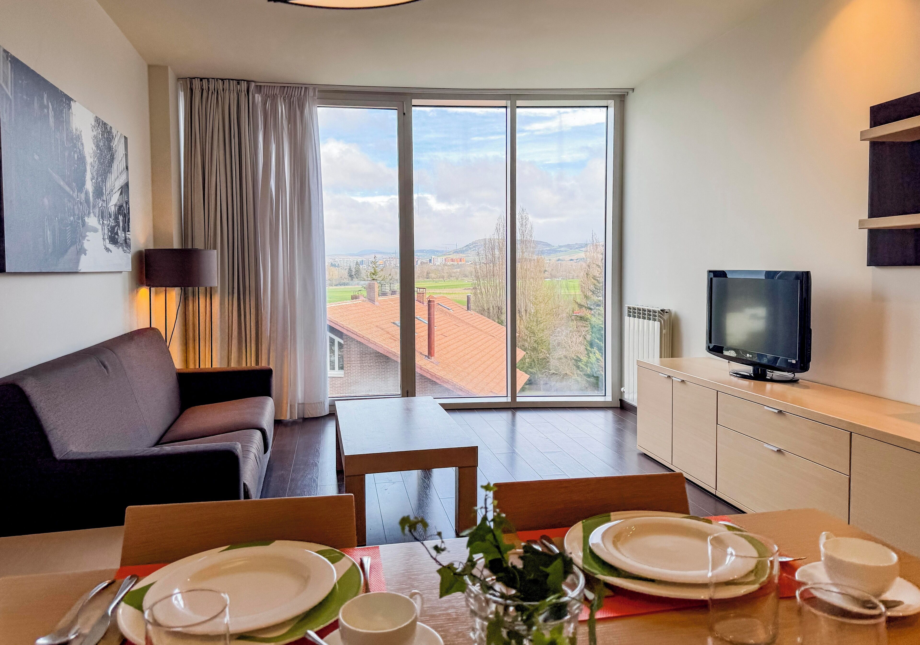 Una sala de estar moderna con un sofá, una mesa de comedor para cuatro personas, un televisor y un ventanal con vistas a la ciudad.