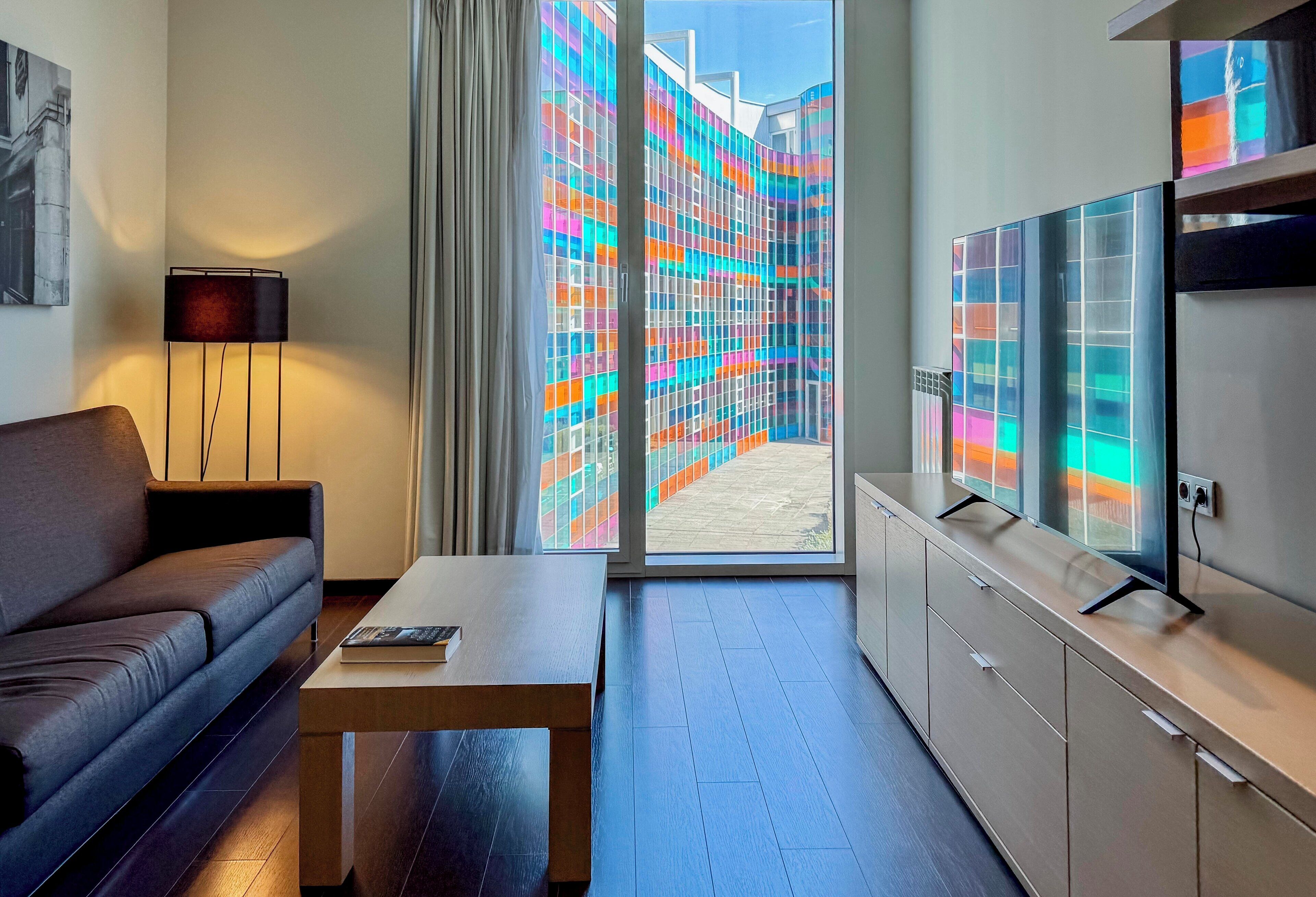 Ett modernt hotellrum med en soffa, ett soffbord, en TV och utsikt över färgglada byggnader med glasfasader utanför.