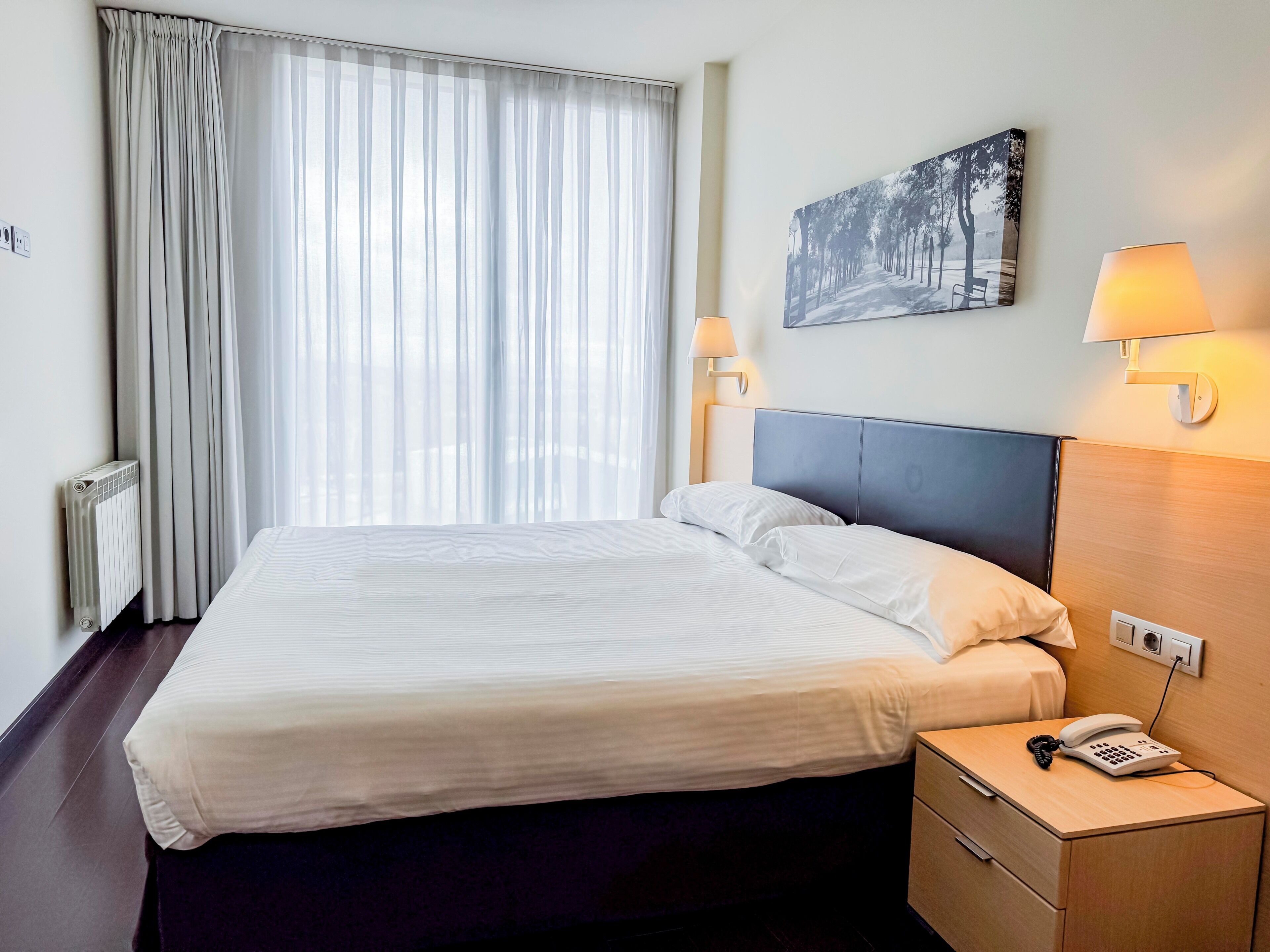 A hotel room with a bed, bedside lamps, a painting, and a phone on the nightstand.