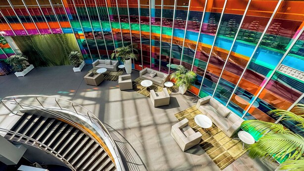 Un atrio moderno con una zona de estar central, una escalera y un muro de cristal con reflejos coloridos.
