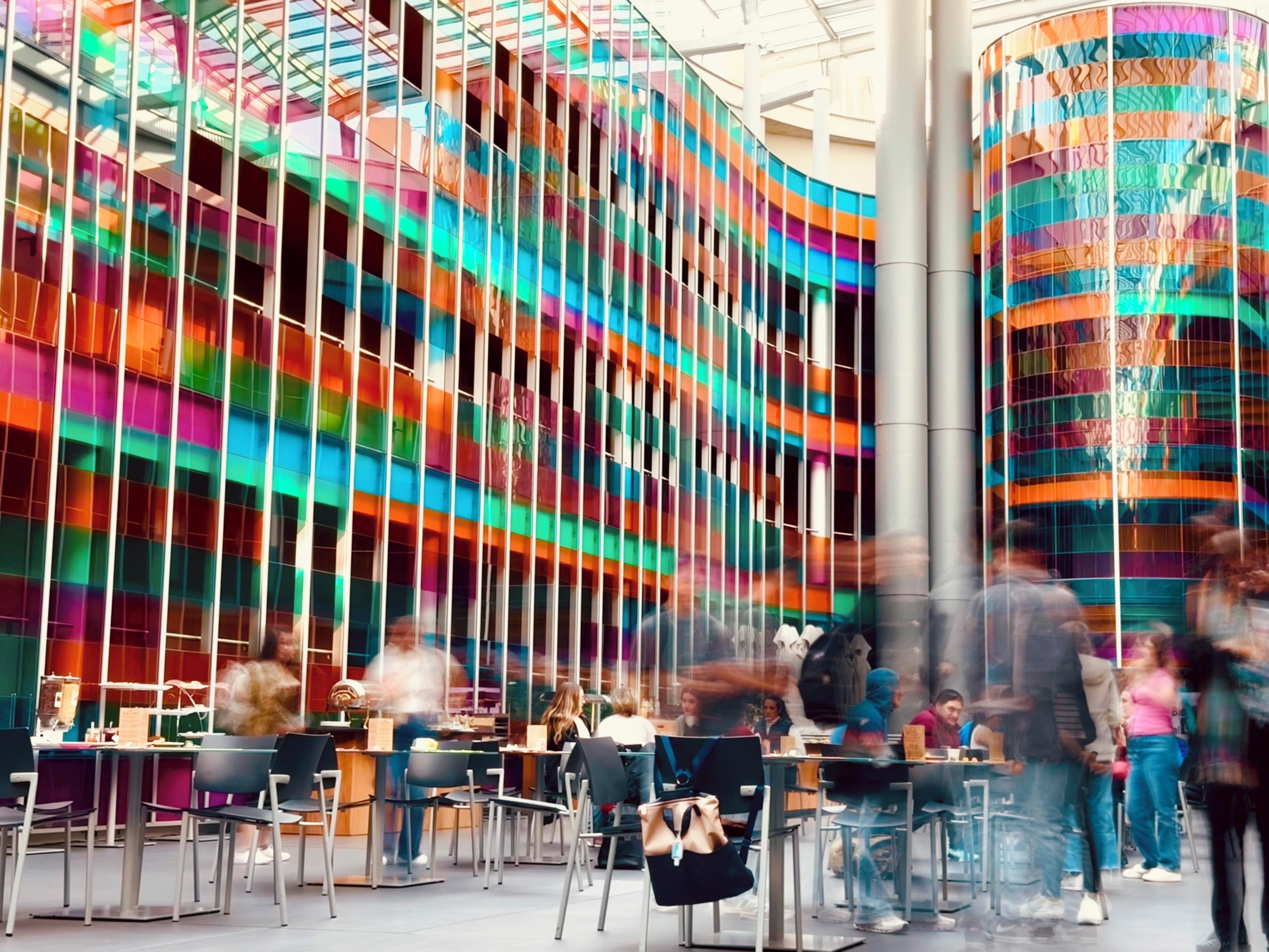A modern indoor space with a colorful, wavy glass facade and a seating area with tables and chairs.
