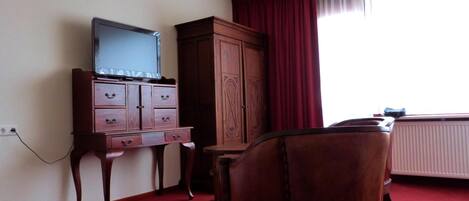 Une chambre d’hôtel avec un meuble en bois, un téléviseur posé dessus, une moquette rouge et une fenêtre avec des rideaux.