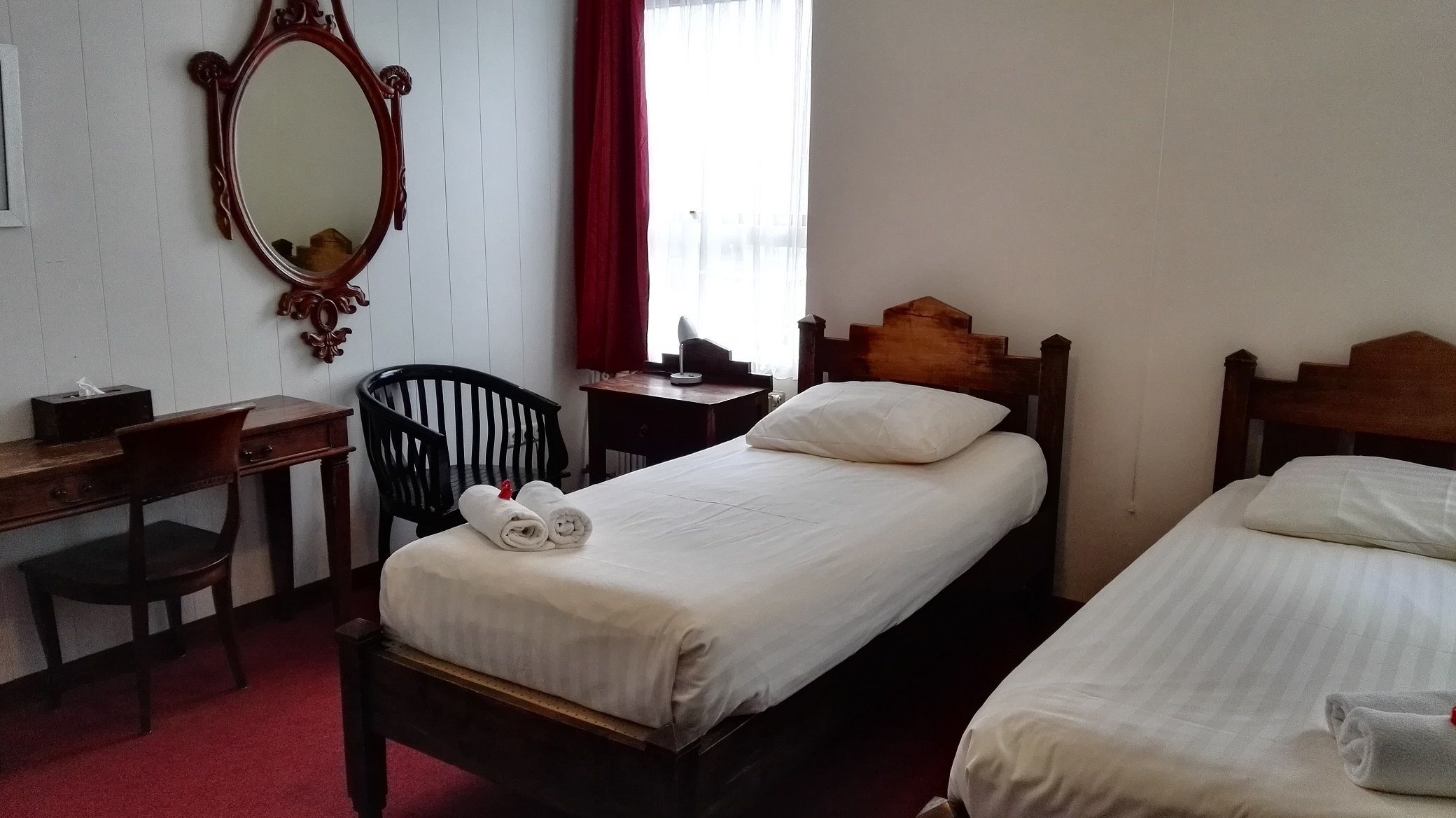 A hotel room with two single beds, a wooden desk, a chair, a mirror, and a window with curtains.