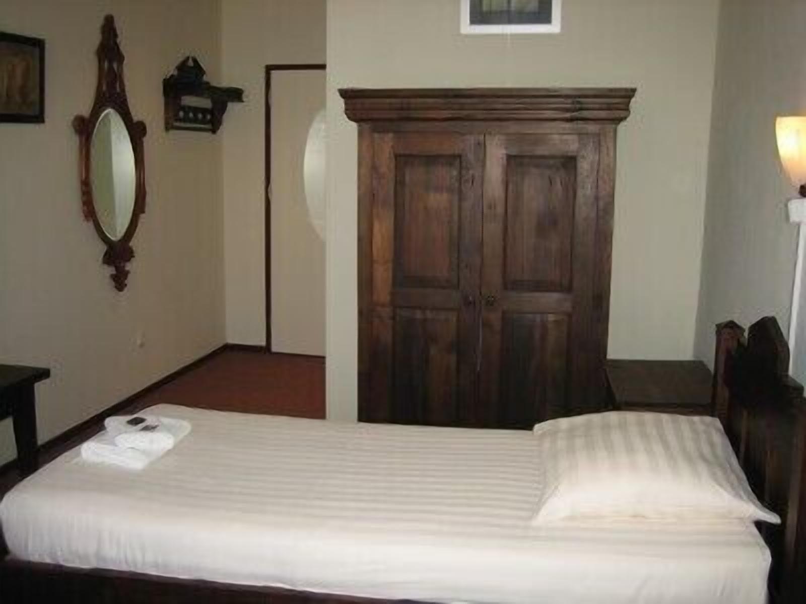 A single bed with a white mattress and pillows, a wooden wardrobe, a mirror, and a wall-mounted light fixture.