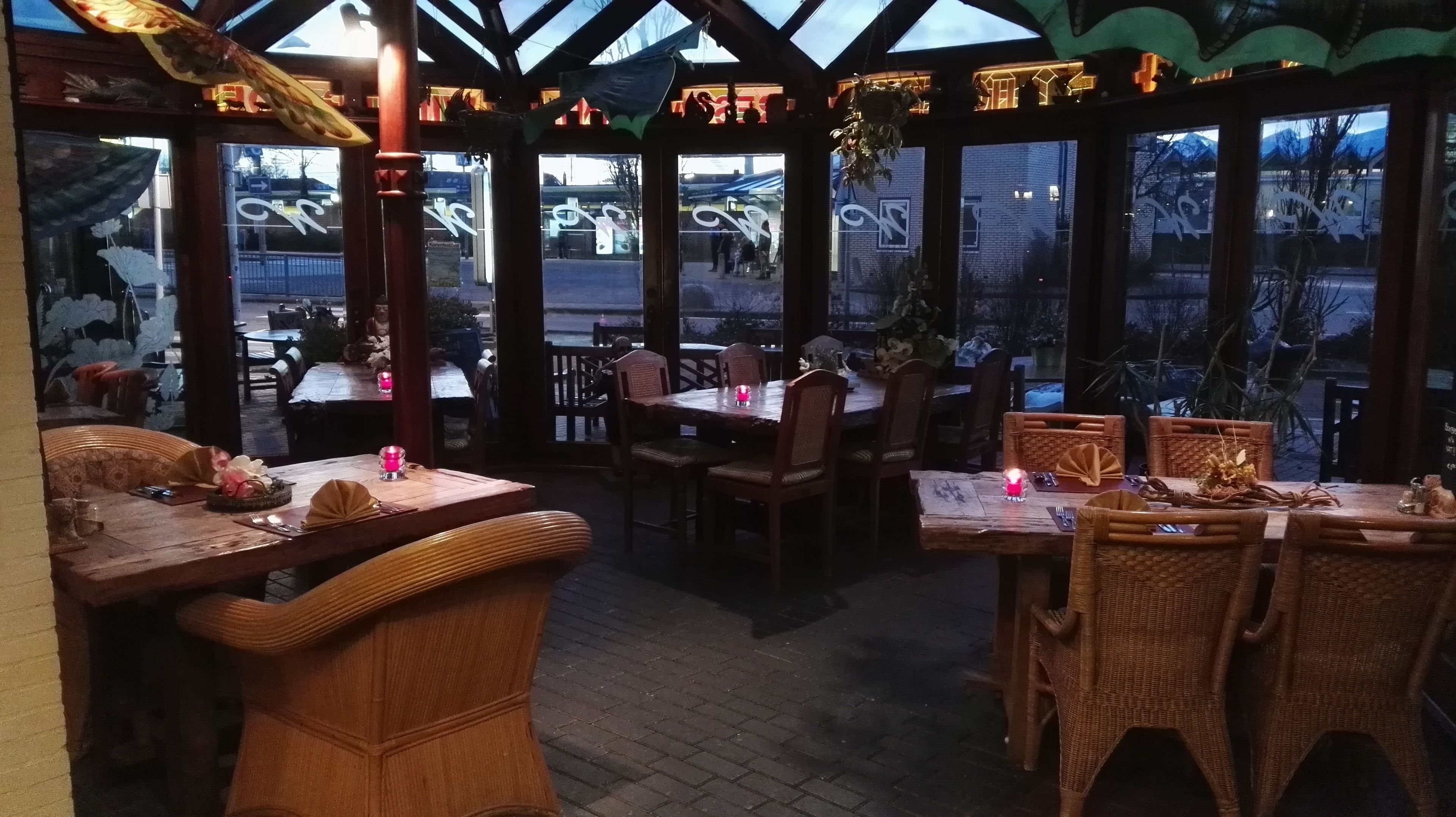 Un restaurante con muebles de madera y ventanas amplias. Las mesas están preparadas con velas y platos.