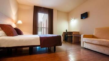 A hotel room with a bed, a desk, a chair, a sofa, a TV, and a window with curtains.
