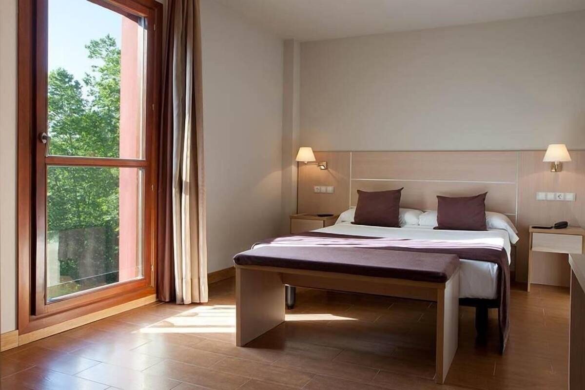 A modern hotel room with a large bed, bedside tables, and a view through a window.