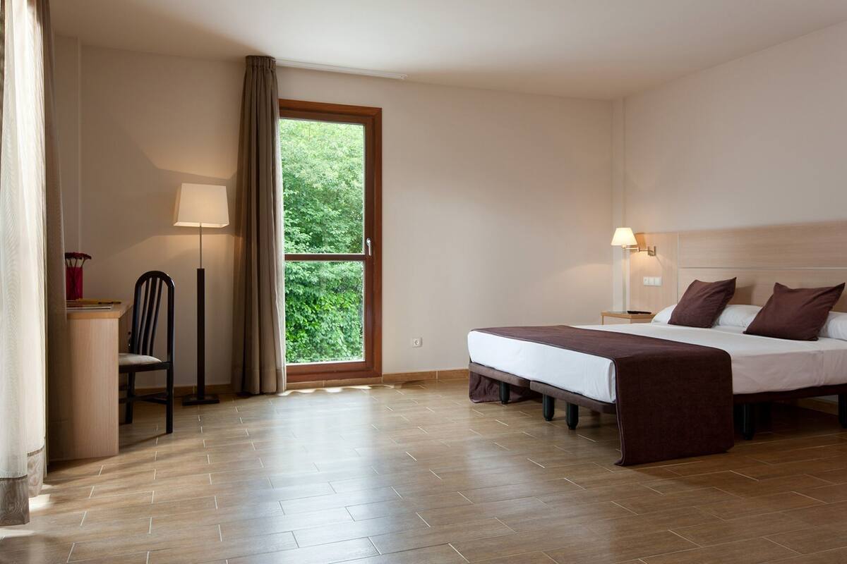 A hotel room with a bed, a desk, a chair, a lamp, and a window with a view of greenery.