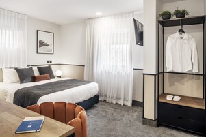 A hotel room with a neatly made bed, a wooden table with a book, a brown chair, a black and white patterned carpet, a white curtain, a framed picture on the wall, and a clothing rack with shoes.