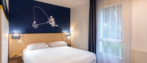Una habitación de hotel con una cama, una obra de arte en la pared que representa a un pescador, dos lámparas de noche, una ventana con cortinas y un pequeño taburete amarillo.