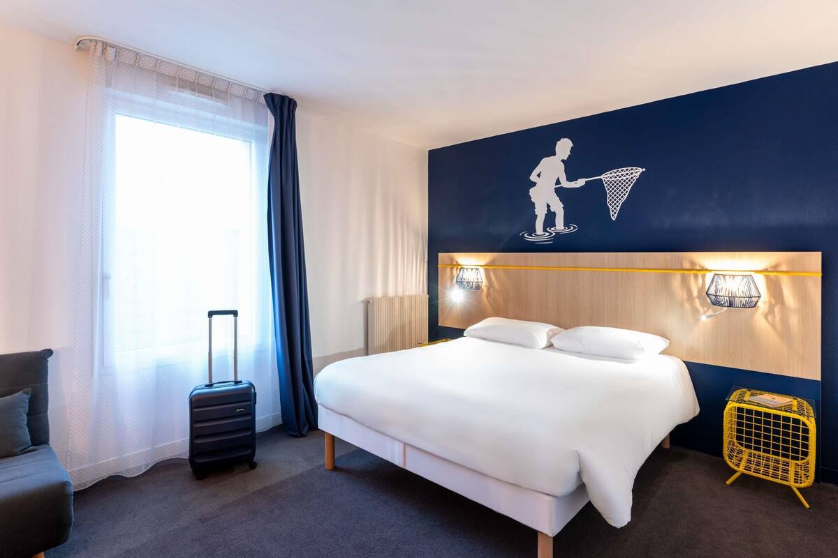 A hotel room with a bed, a suitcase, a chair, a wall-mounted lamp, and a decorative wall piece.