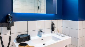 A modern bathroom with a white sink, a handheld hair dryer, a black bag, a wooden brush, and a bottle of shampoo.