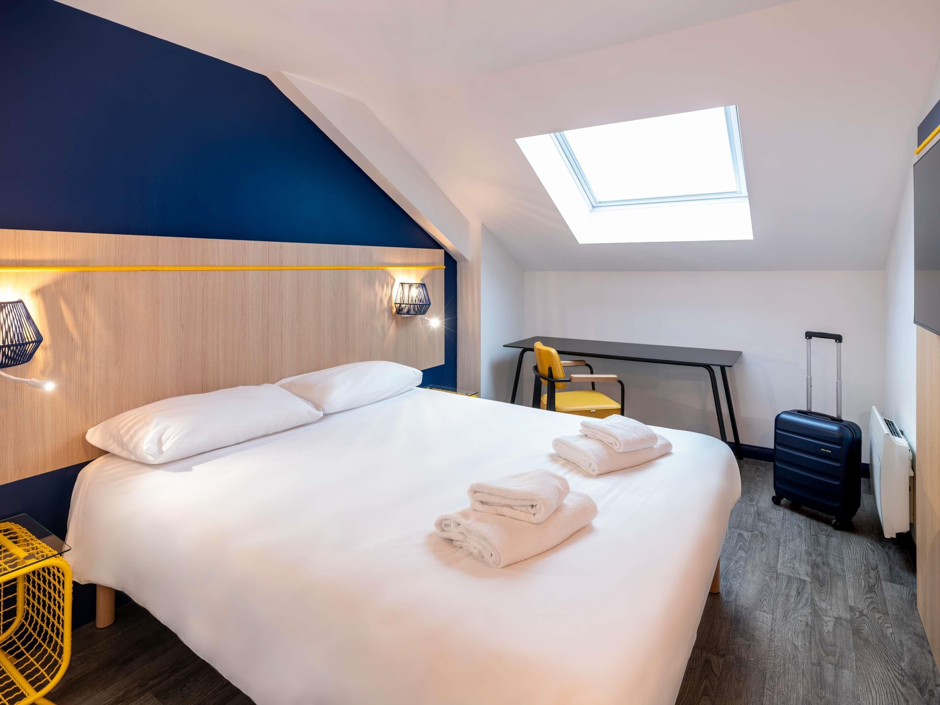 A modern hotel room with a large bed, a desk, a chair, and a skylight.