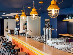 A bar area with multiple hanging yellow pendant lights, a bar counter with glasses and bottles, and a chalkboard menu in the background. - ibis Styles Morlaix (Morlaix)