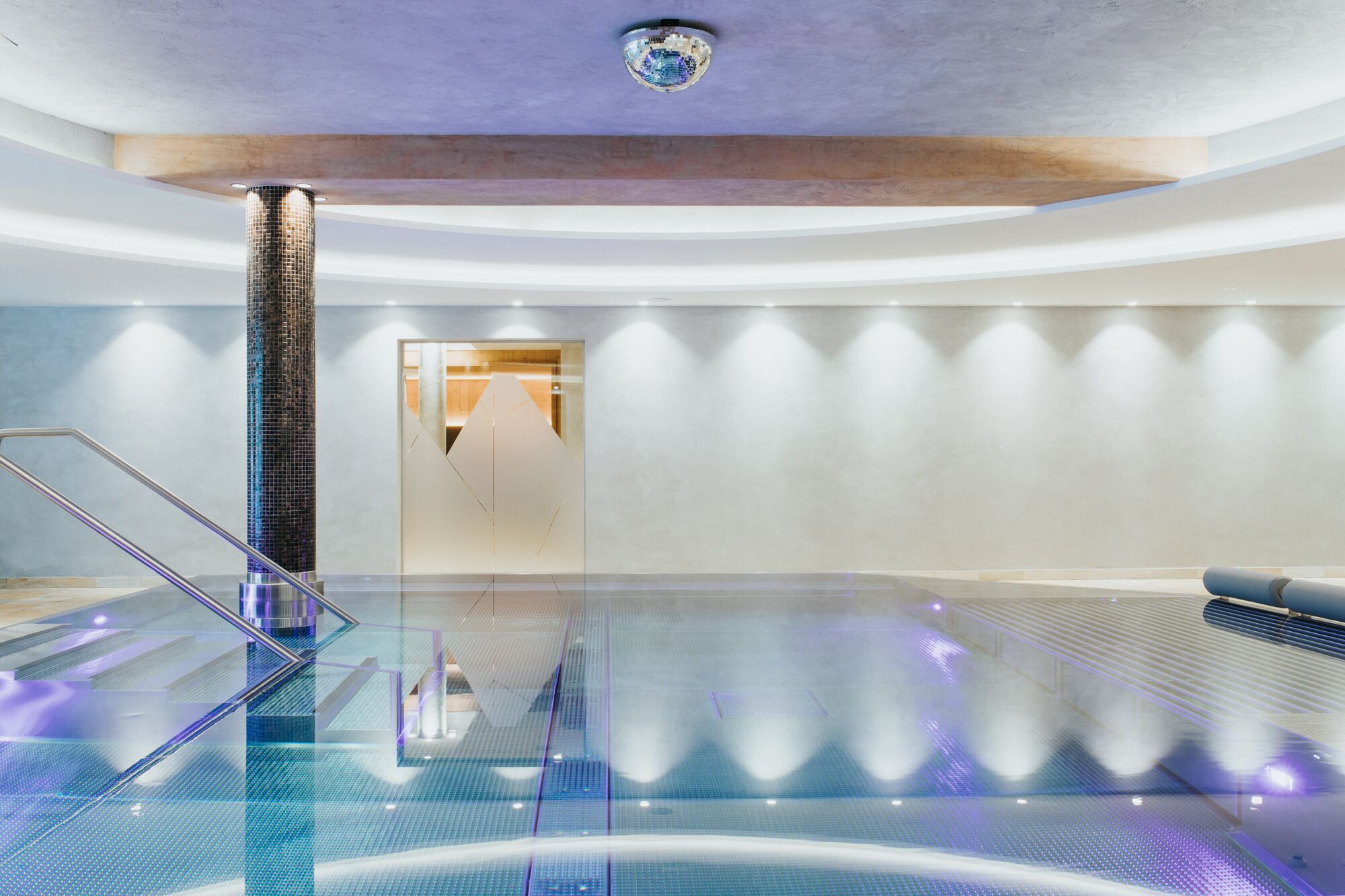 Una piscina interior moderna con suelo de vidrio, una columna central y un diseño minimalista.