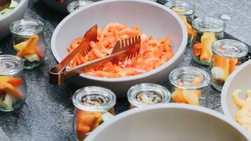 A buffet table with various dishes including a large bowl of shredded carrots, a bowl of cherry tomatoes, and several small jars of pickled vegetables.