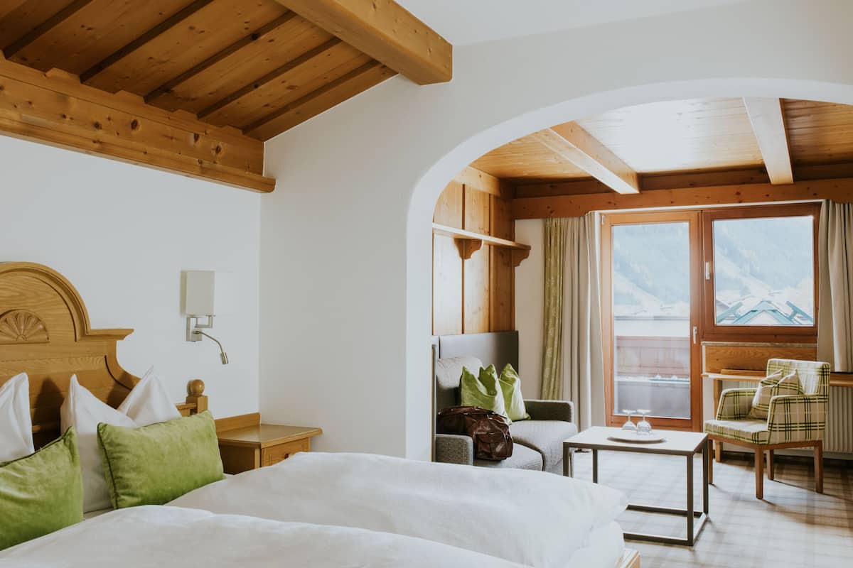 Een hotelkamer met een bed, een houten hoofdbord, een tafeltje, een stoel en uitzicht op de bergen door een groot raam.