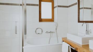 A modern bathroom with a white tiled wall, a wooden-framed window, a bathtub, a sink with a mirror, and a towel rack.