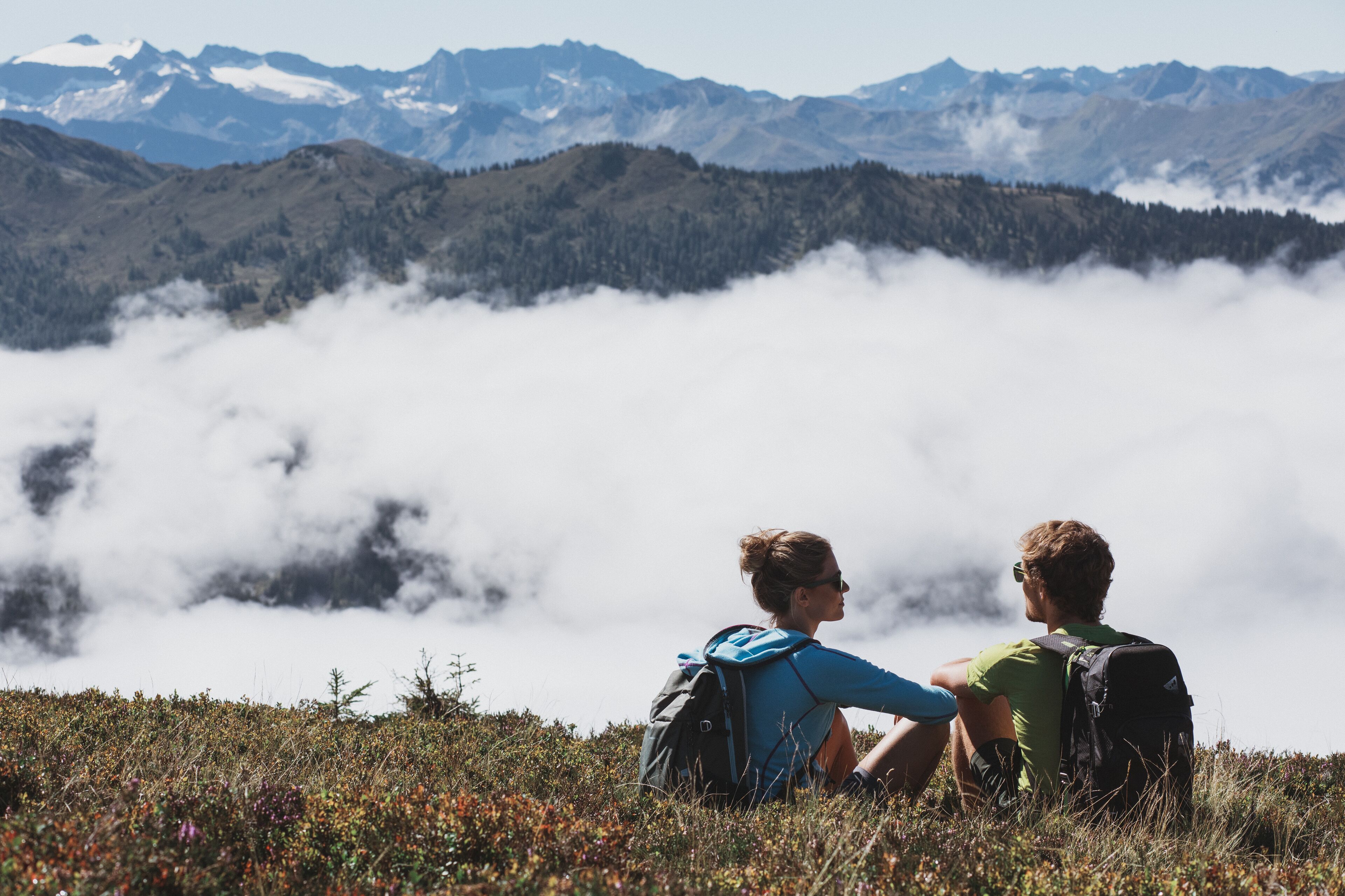 Two people sitting on a grassy hill with a mountainous backdrop.