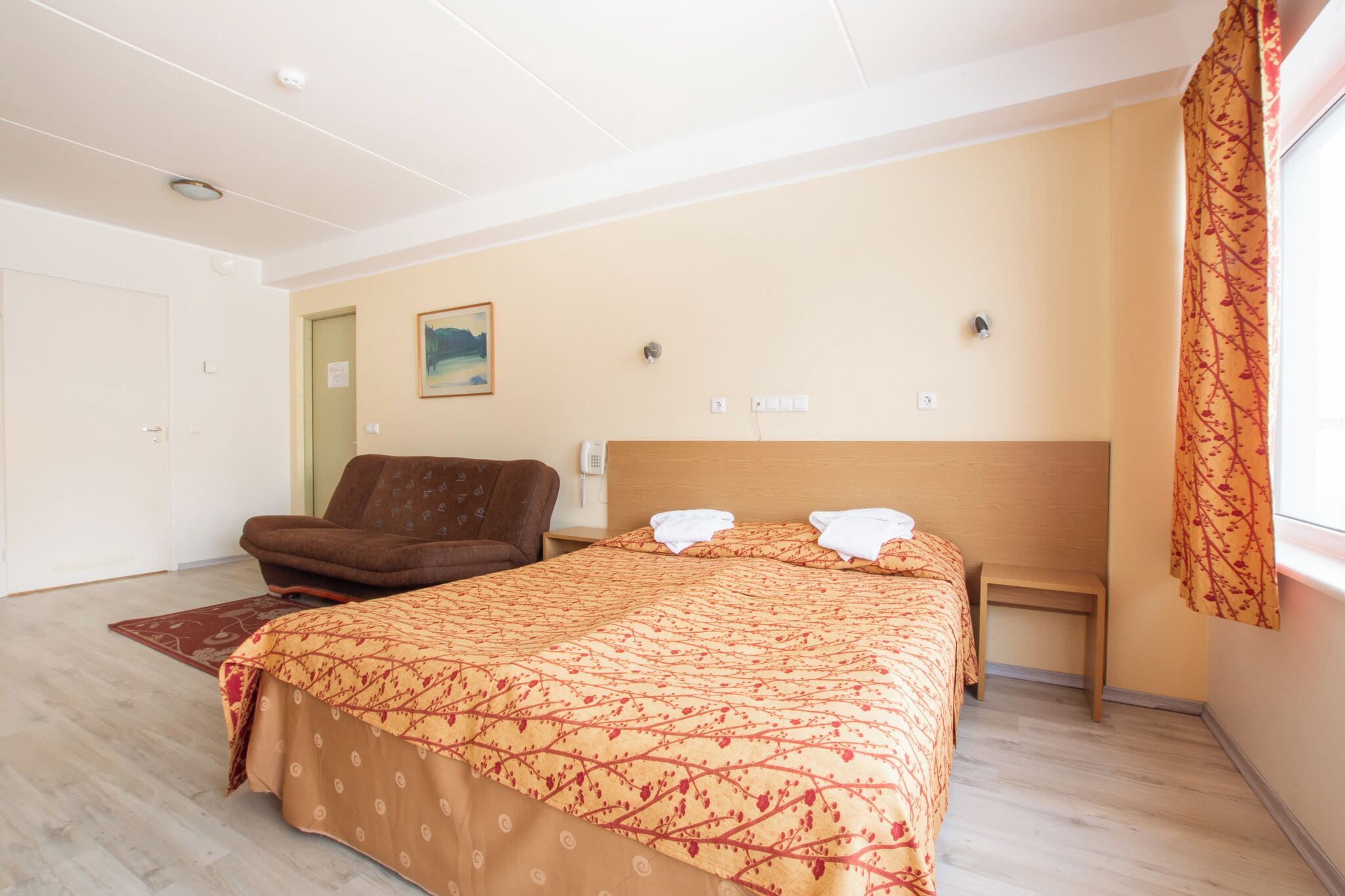 A hotel room with a bed, a chair, a small table, and a window with curtains.