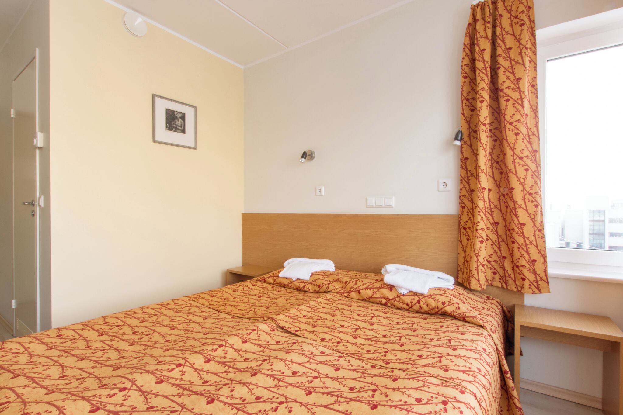 A hotel room with a bed, a bedside table, a window with curtains, a wall-mounted light, and a framed picture.