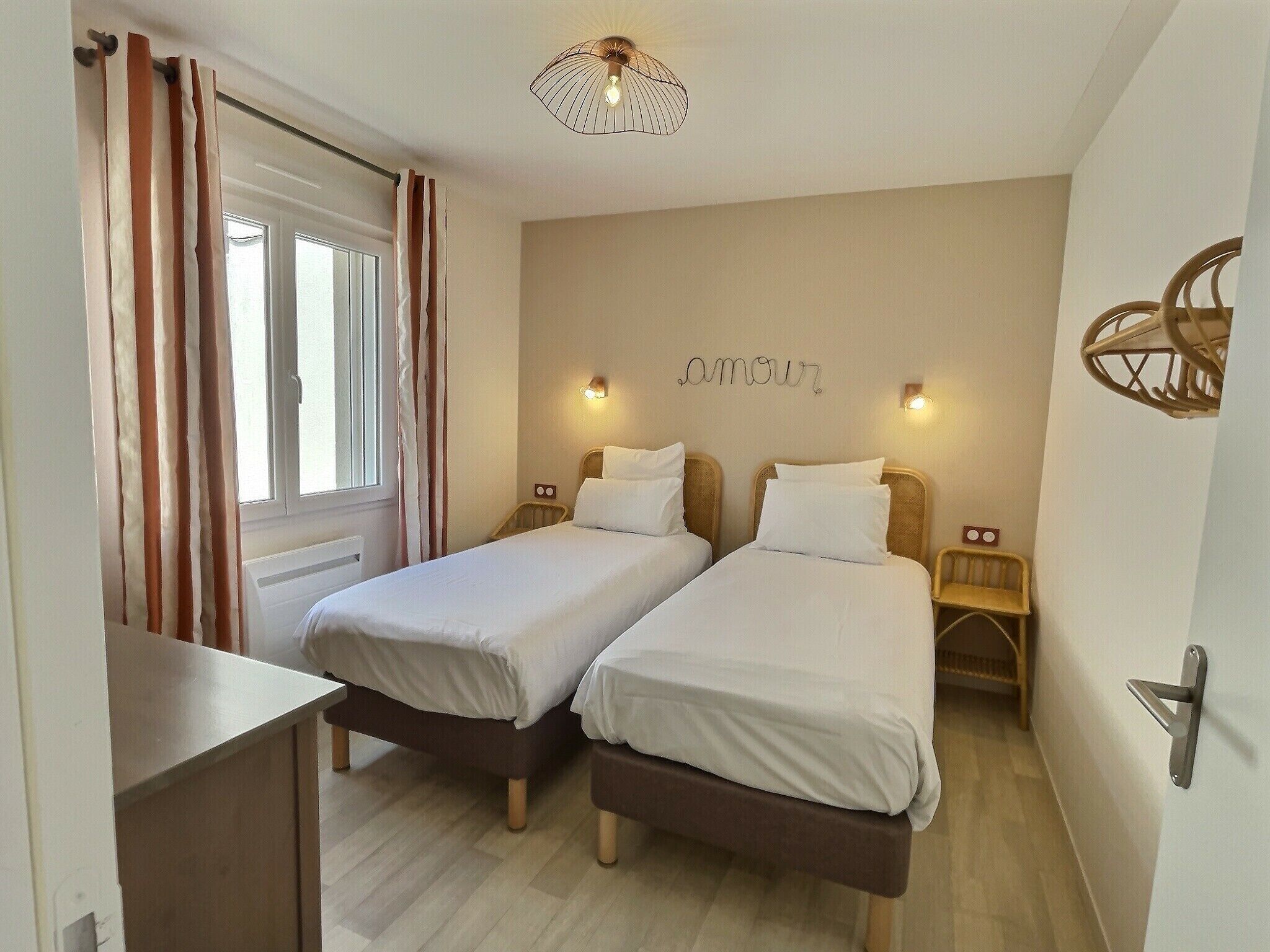 A double room with two beds, a window with curtains, a wall-mounted light, and a ceiling light fixture.