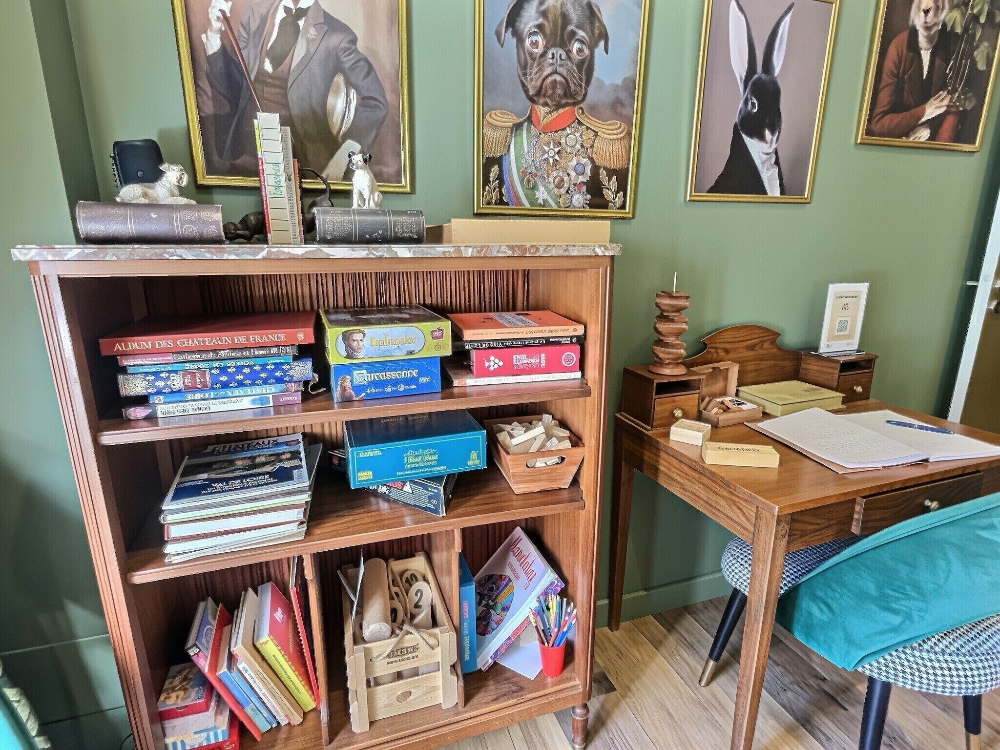 A room with a wooden bookshelf filled with books and board games, a desk with papers and a pen, and a chair with a teal cushion.